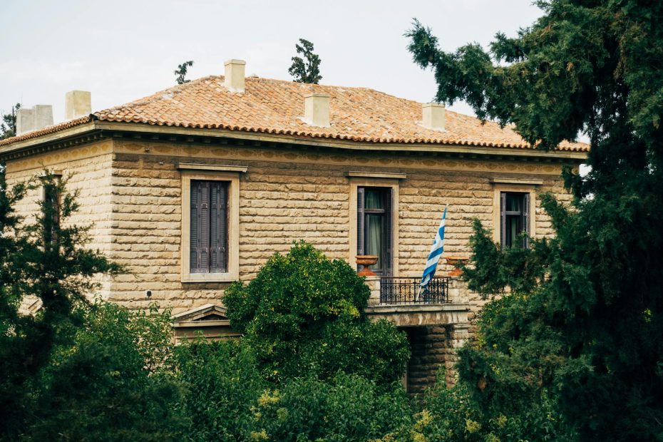 Historic stone villa with a Greek flag surrounded by greenery, showcasing classic architecture and lush landscape.